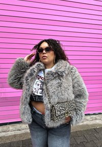Giacca in pelliccia sintetica grigia con trama spessa, abbinata a una maglietta grafica, jeans grigi a vita alta e una borsa con stampa leopardata. Sfondo rosa.