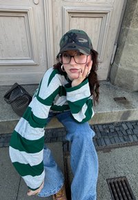 Chemise à manches longues rayée verte et grise, jean bleu évasé, casquette camouflage, lunettes transparentes et sac noir tissé posés sur une surface en pierre.