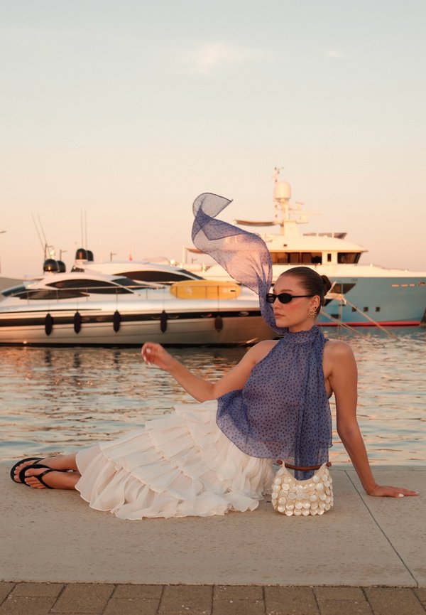 Frau in einem blauen transparenten Top und weißem Rock sitzt am Wasser mit Yachten im Hintergrund während des Sonnenuntergangs.