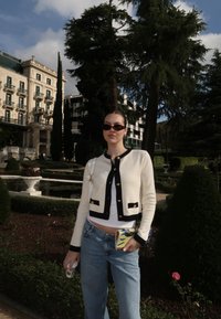 En kremfarget cardigan med svarte detaljer og gullknapper, kombinert med lyseblå jeans. Bakgrunnen inkluderer grøntområder og en bygning.