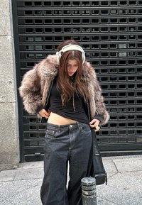 Jeune femme portant des écouteurs et une veste en fourrure ajuste son jean noir ample alors qu'elle se tient sur un trottoir en ville devant une grille en métal.