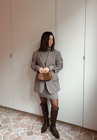 Mujer con blazer gris y vestido, sosteniendo un bolso marrón tejido, de pie sobre un suelo de baldosas frente a una pared blanca texturizada.