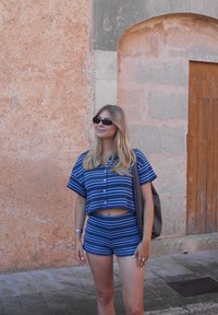 Niebiesko-biała, w paski krótka bluzka z krótkim rękawem i pasujące szorty, wykonane z lekkiej tkaniny. Osoba niesie brązową, teksturowaną torbę.