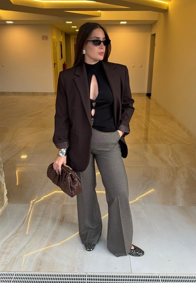 Blazer marrón oversize, top negro con cut-out, pantalones grises de talle alto, clutch marrón tejido, bailarinas negras con tachuelas. Fondo de suelo liso.