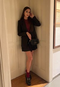 Μαύρο ταγέρ σακάκι πάνω από ένα μπορντό τοπ, συνδυασμένο με μια σκούρα φούστα. Τα αξεσουάρ περιλαμβάνουν μια μικρή μαύρη υφασμάτινη τσάντα και σκούρες λόφερ με χρυσές λεπτομέρειες.