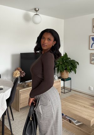 Mujer con cabello oscuro y ondulado, vestida con una blusa marrón de manga larga y una falda gris de cuadros, sosteniendo un bolso negro en una sala de estar moderna.