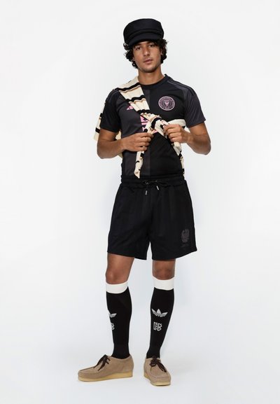 Camiseta deportiva negra con logo, bufanda a rayas en crema y marrón, pantalones cortos negros, calcetas negras hasta la rodilla y zapatos marrón claro.