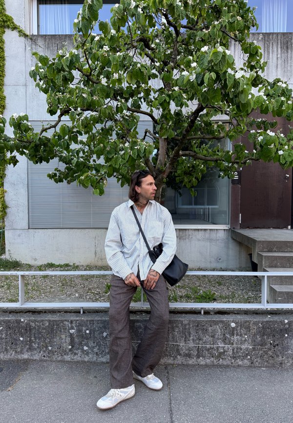Homme aux cheveux longs, en chemise rayée et pantalon marron, assis sur un rebord sous un arbre feuillu en fleurs, avec un bâtiment en arrière-plan.