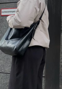 Isik, kes kannab heledat jope ja musti pükse, hoiab suurt musta nahast õlakotti ja seisab seina ääres, kus on saksa keeles silt.