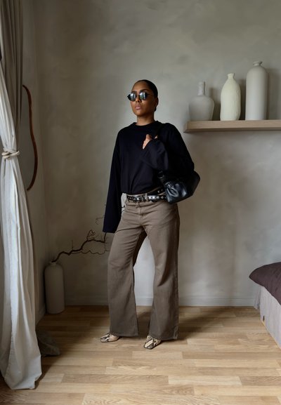 Mujer con gafas de sol, camisa negra de manga larga, pantalones anchos marrones, zapatos estampados y llevando un bolso negro, parada en el interior cerca de jarrones.