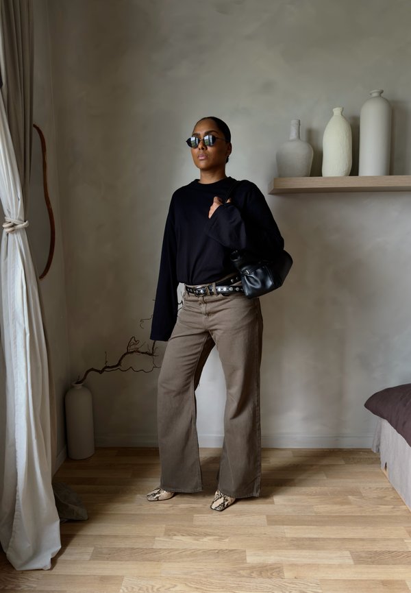 Ženska z sončnimi očali, črno majico z dolgimi rokavi, širokimi rjavimi hlačami, vzorčastimi čevlji in z rdečo torbico, stoji v notranjosti blizu vaz.