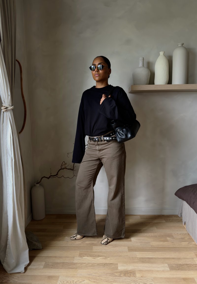 Naine päikeseprillidega, must pika varrukaga särk, laiad pruunid püksid, mustriga kingad ja must käekott ning seisab toas vaaside lähedal.