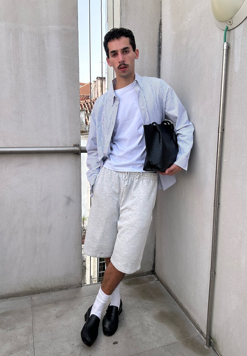 Homme appuyé contre un mur portant un T-shirt blanc, une chemise rayée boutonnée, un short gris, des chaussettes blanches, des chaussures noires, tenant un sac tissé noir.