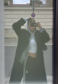 Persona con felpa grigia sotto un cappotto nero che scatta un selfie attraverso una superficie riflettente coperta da una rete, con un cartello di divieto di sosta sullo sfondo.