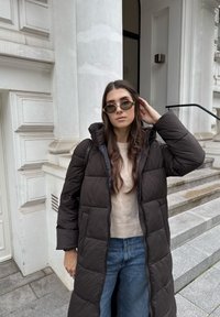 Pikk must puffer-kurjused, millel on kapuuts, tilgutusmuster, koos beežist kampsunist ja sinistest teksadest. Ümmargused rohelised päikeseprillid. Kivised astmed taustal.