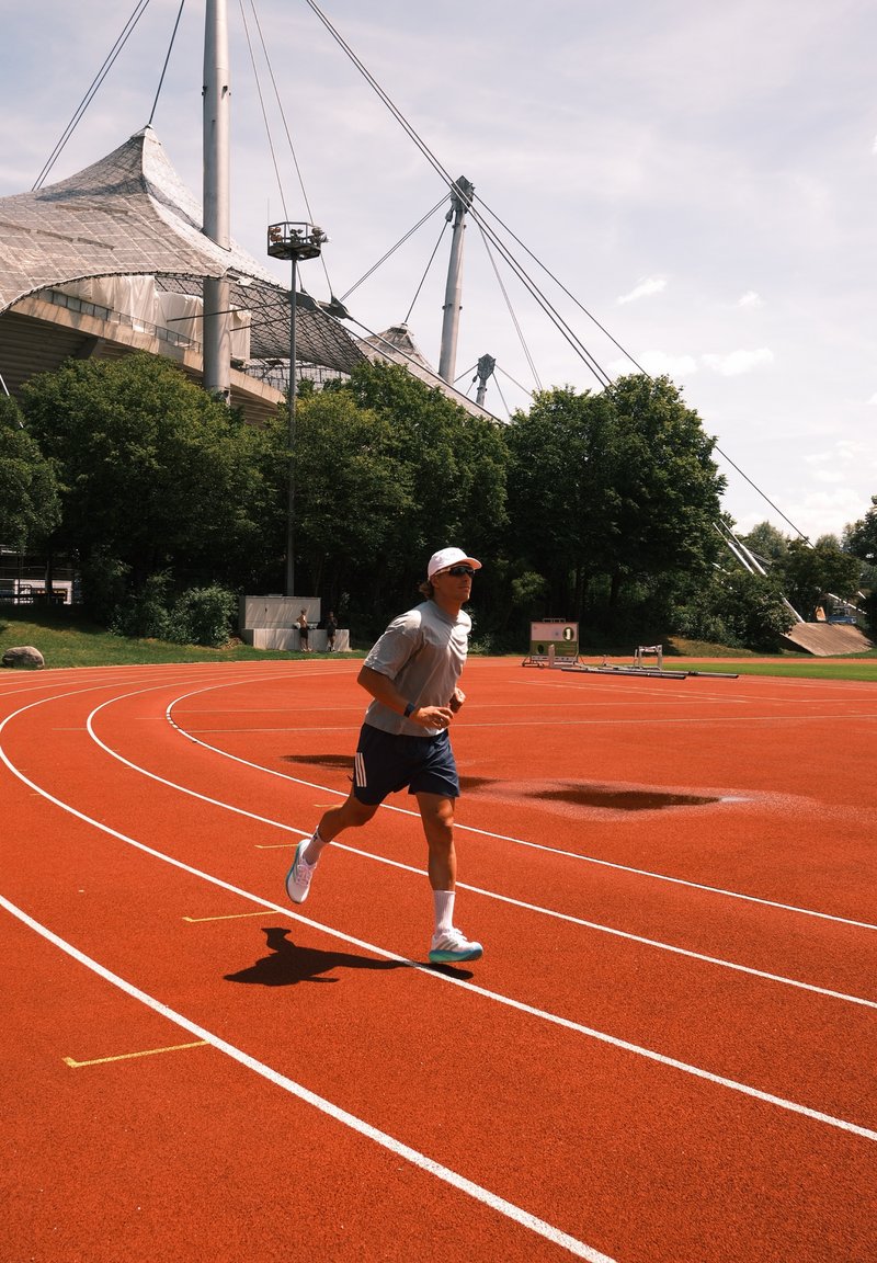Mand iført kasket og solbriller, der jogger på en rød løbebane med træer og en stor stadionstruktur i baggrunden.