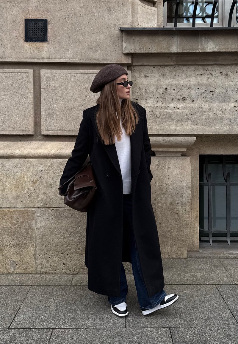 Casaco preto de abotoamento duplo, suéter branco, jeans escuros. Boina castanha e óculos de sol. Mala castanha de ombro, ténis pretos e brancos. Fundo de parede de pedra.