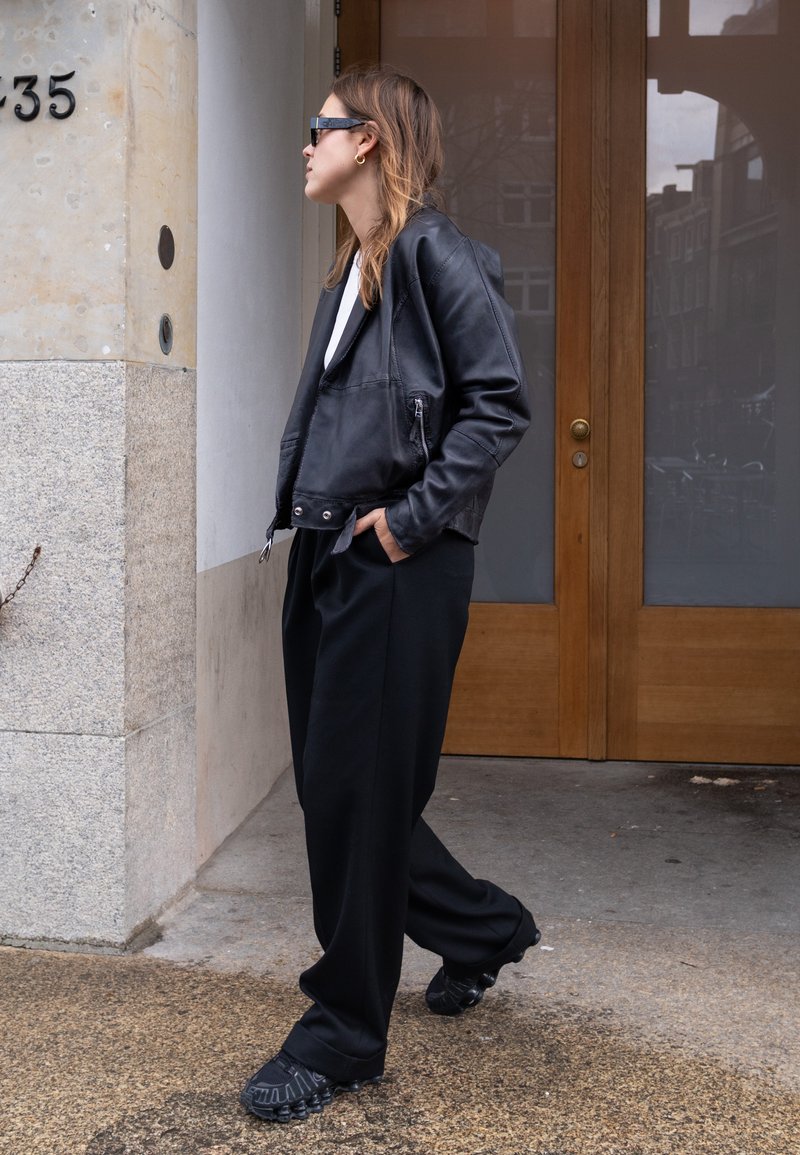 Henkilö, joka on pukeutunut mustaan nahkatakkiin, mustiin housuihin ja mustiin lenkkareihin, seisoo rakennuksen sisäänkäynnin lähellä, jossa on puutavaraa olevat kaksiosaiset ovet.