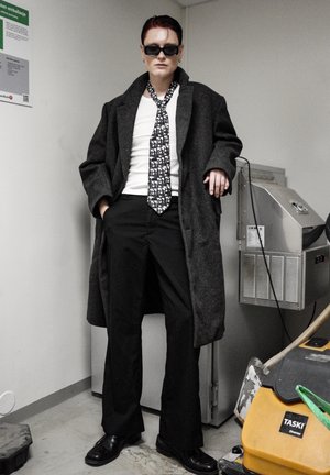 Man wearing a black overcoat, white shirt, patterned tie, and black flared trousers. Accessories include black sunglasses and black shoes.