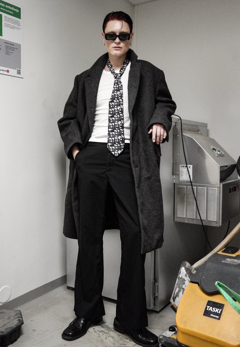 Homme portant un manteau noir, une chemise blanche, une cravate à motifs et un pantalon noir évasé. Les accessoires incluent des lunettes de soleil noires et des chaussures noires.