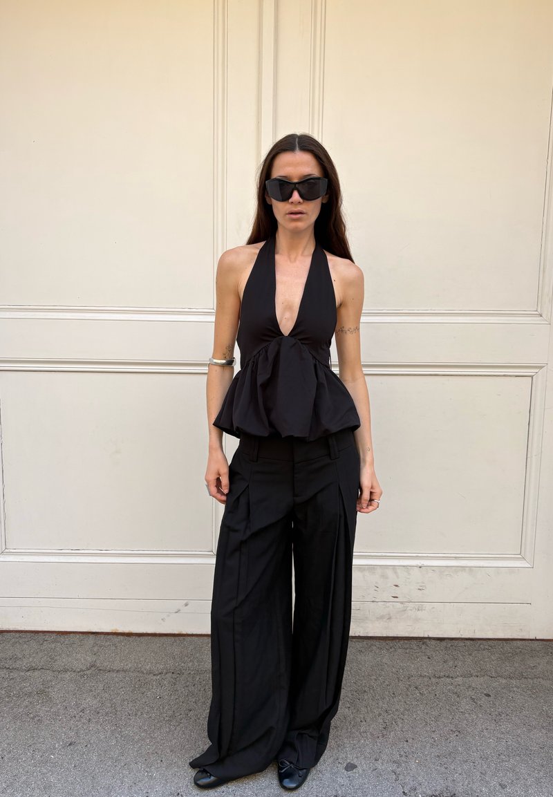 Femeie cu păr lung și închis la culoare, purtând ochelari de soare negri supradimensionați, un top negru fără mâneci cu guler halter, pantaloni negri largi și pantofi negri.