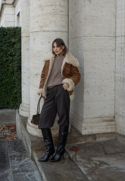 Chaqueta de piel vuelta marrón con forro crema, suéter beige de canalé, pantalones oscuros cortos y botas negras de tacón fino hasta la rodilla, sosteniendo un bolso pequeño.