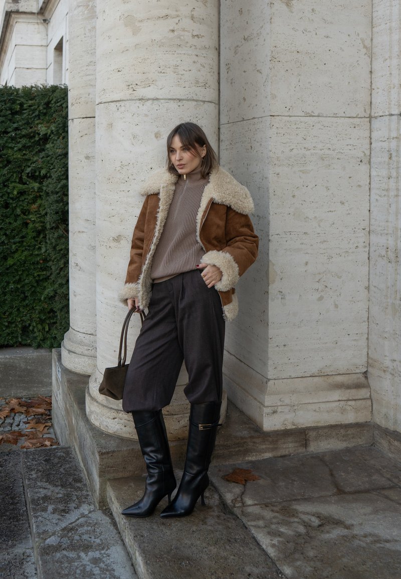 Veste marron en peau retournée avec doublure crème, pull côtelé beige, pantalon court foncé et bottes noires à talons aiguilles montant jusqu'aux genoux, tenant un petit sac.