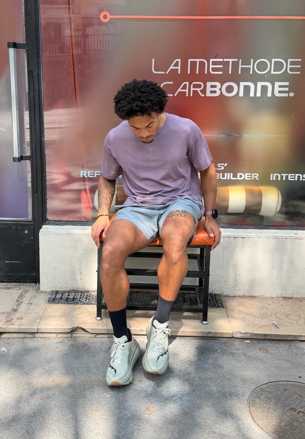 Mann in lila Hemd und grauer Shorts sitzt auf einer orangenen Bank vor einem Laden mit der Aufschrift „La Méthode Carbonne“ auf dem Fenster hinter ihm.