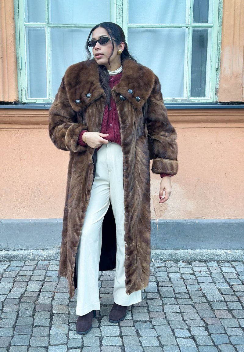 Femme portant un long manteau en fourrure brune, un pull bordeaux, un pantalon blanc, des bottes à talons noires, des lunettes de soleil, debout dans une rue pavée près d'un bâtiment avec fenêtres.