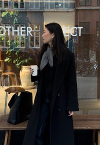 Manteau noir croisé à double boutonnage, écharpe grise, tenant une tasse de café, avec un jean foncé et un sac noir ; en arrière-plan, des plantes et du mobilier en bois.