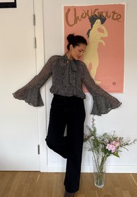 Schwarz-weiß gepunktete Bluse mit Trompetenärmeln, hohem Halsausschnitt und gerafftem Saum, kombiniert mit schwarzen Hosen. Blumen in einer klaren Vase in der Nähe.