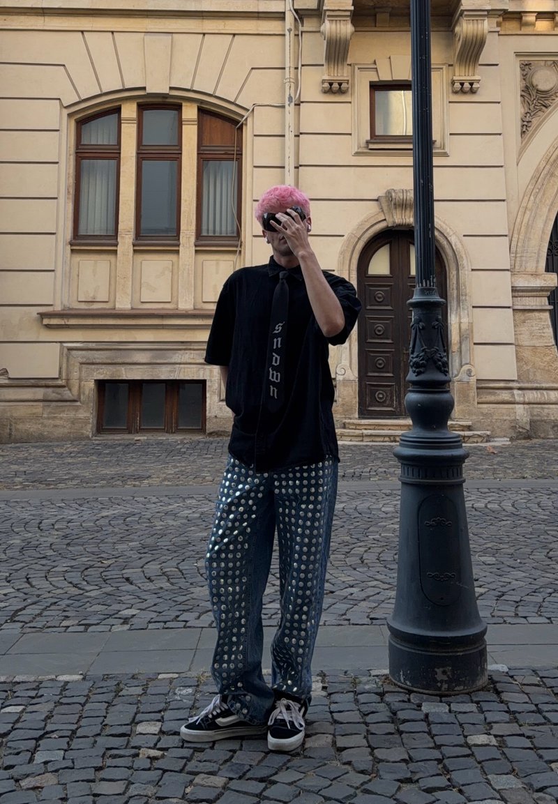 Personne aux cheveux roses portant une chemise noire et un pantalon à motifs, se tient dans une rue pavée à côté d’un lampadaire, couvrant son visage avec sa main.