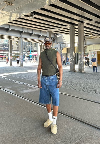 Hombre vestido con una camiseta sin mangas verde de punto, pantalones cortos de mezclilla largos, calcetines blancos, zapatos beige sin cordones, gafas de sol y una gorra, de pie bajo un puente.