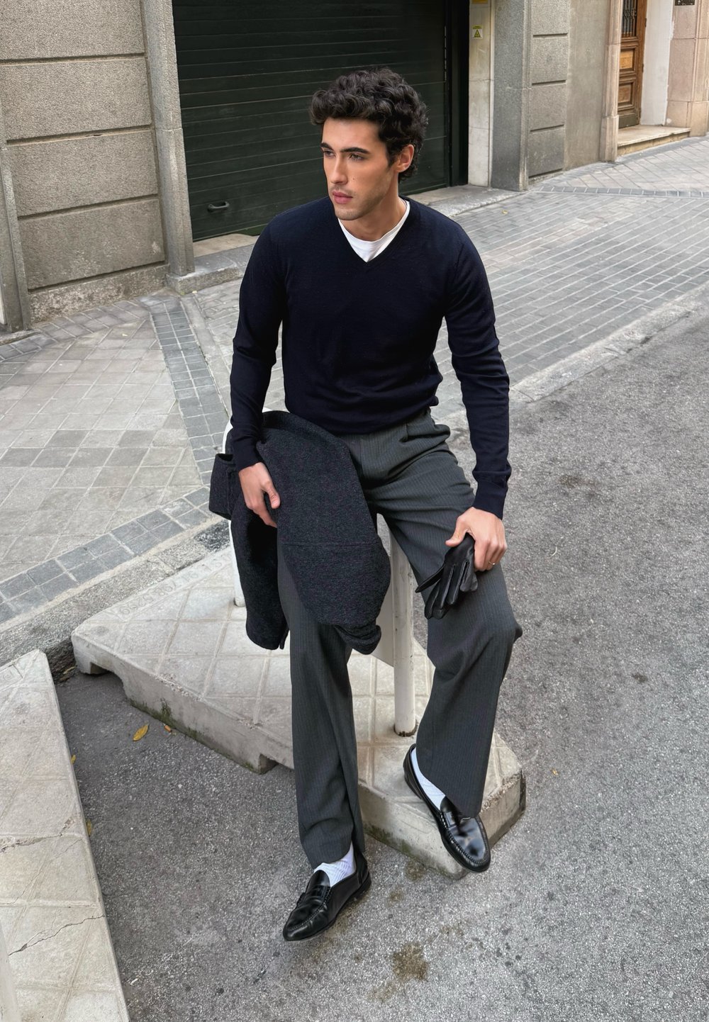 Maglione a scollo a V navy, pantaloni grigi a righe sottili, mocassini neri, che tiene un cappotto scuro e guanti neri; in piedi su un blocco di cemento in un ambiente urbano.
