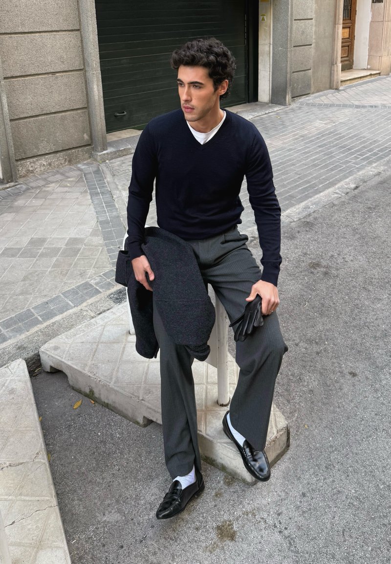 Pull à col en V bleu marine, pantalon gris à fines rayures, mocassins noirs, tenant un manteau foncé et des gants noirs ; debout sur un bloc de béton dans un cadre urbain.