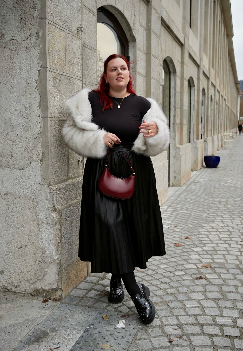 Μαύρη πλισέ φούστα, μαύρο μπλουζάκι και λευκό ψεύτικο γούνινο παλτό. Μπορντό τσάντα χειρός και μαύρα πλατφόρμα παπούτσια με καρφιά. Φόντο με cobblestone δρόμο.
