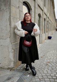 Μαύρη πλισέ φούστα, μαύρο μπλουζάκι και λευκό ψεύτικο γούνινο παλτό. Μπορντό τσάντα χειρός και μαύρα πλατφόρμα παπούτσια με καρφιά. Φόντο με cobblestone δρόμο.