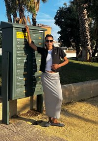 Μαύρο κοντομάνικο πουκάμισο, λευκό t-shirt από κάτω, γκρι φούστα και μαύρα και λευκά παπούτσια με σχέδια. Στέκει κοντά σε πράσινη μονάδα ταχυδρομείου.