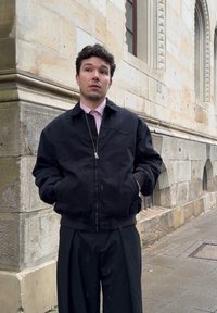 A young man with curly hair, dressed in a black jacket and trousers, stands with his hands in his pockets against a stone building on a wet pavement.