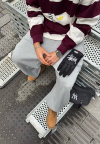 Black gloves with white text, grey sweatpants, maroon and cream striped sweatshirt, tan boots, silver watch, and a dark bag with a logo.