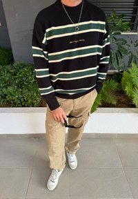Sweater listrado preto e verde com gola canelada, calças cargo bege e ténis brancos. A segurar óculos de sol; ajuste descontraído e tecido texturizado.