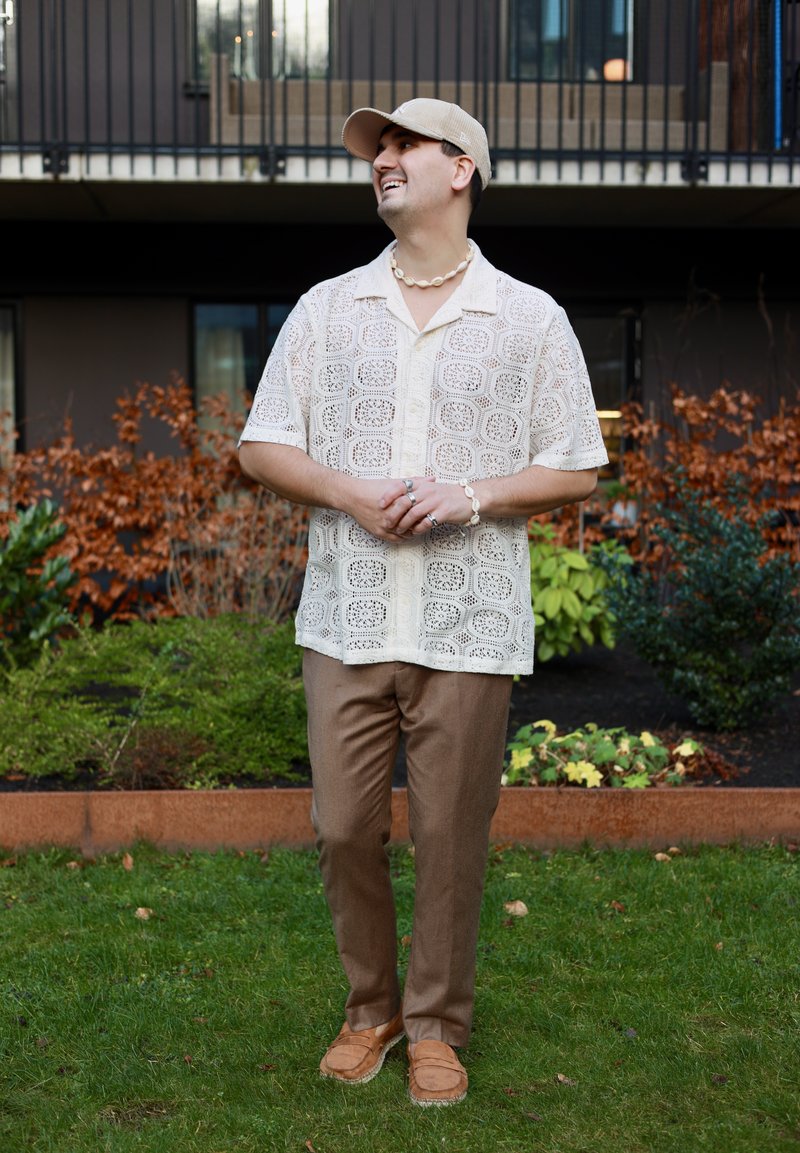 Beige horgolt inget, barna nadrágot és sapkát viselő férfi áll a kertben egy modern épület előtt, mosolyogva, kezét összekulcsolva.