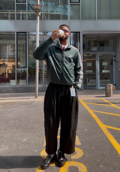 Chaqueta verde con cremallera y rayas blancas, pantalones anchos negros, zapatos negros, sosteniendo una bebida, de pie en una calle con líneas amarillas.