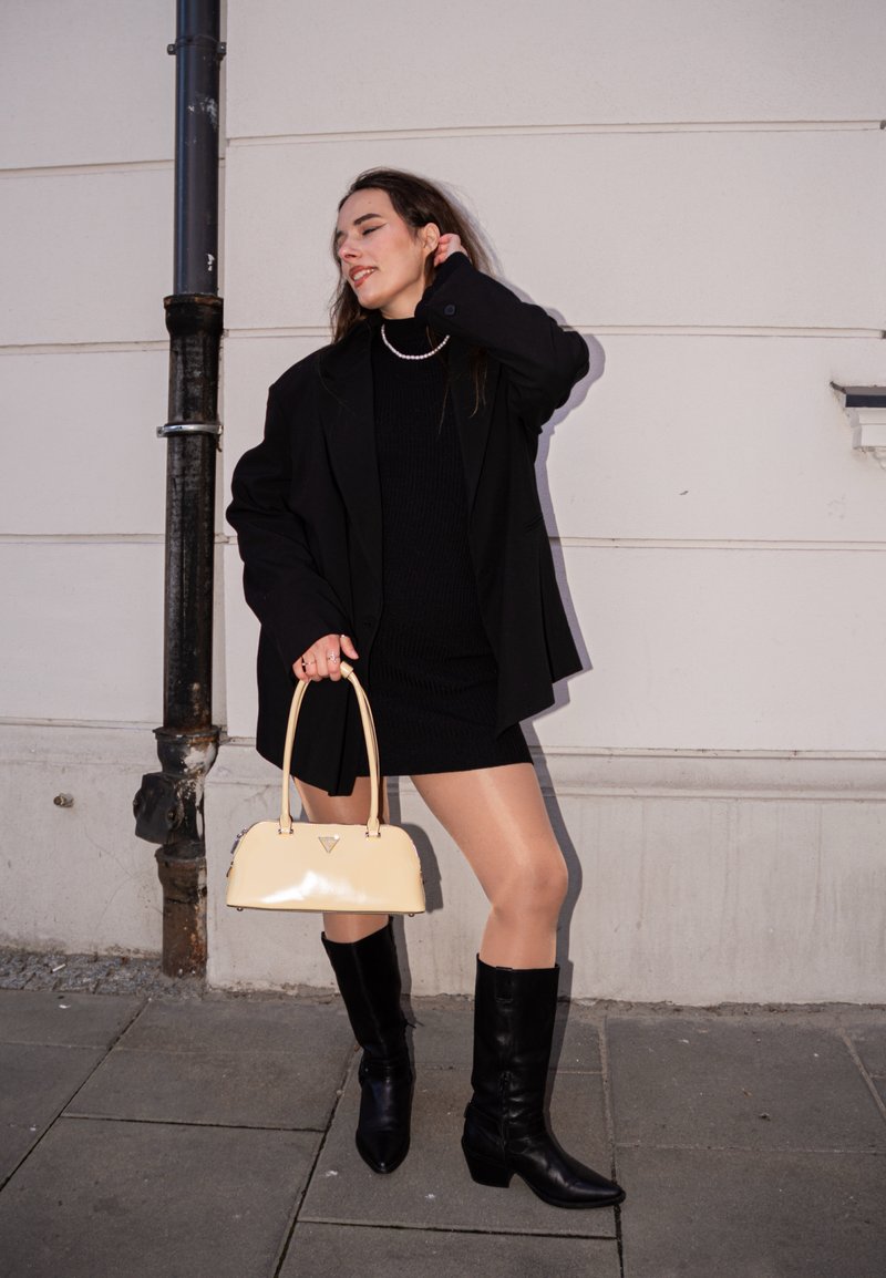 Frau trägt schwarzes Kleid, übergroßen Blazer und kniehohe schwarze Stiefel, hält eine beige Handtasche und steht auf dem Bürgersteig vor einer weißen Wand.