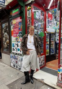 Modelo a usar uma jaqueta oversized castanha, t-shirt branca, calções cinzentos e botas altas pretas, acessorizada com colares dourados em camadas.