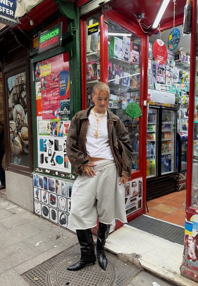 Modelo luciendo una chaqueta oversized marrón, camiseta blanca, pantalones cortos grises y botas negras hasta la rodilla, complementado con cadenas de oro en capas.
