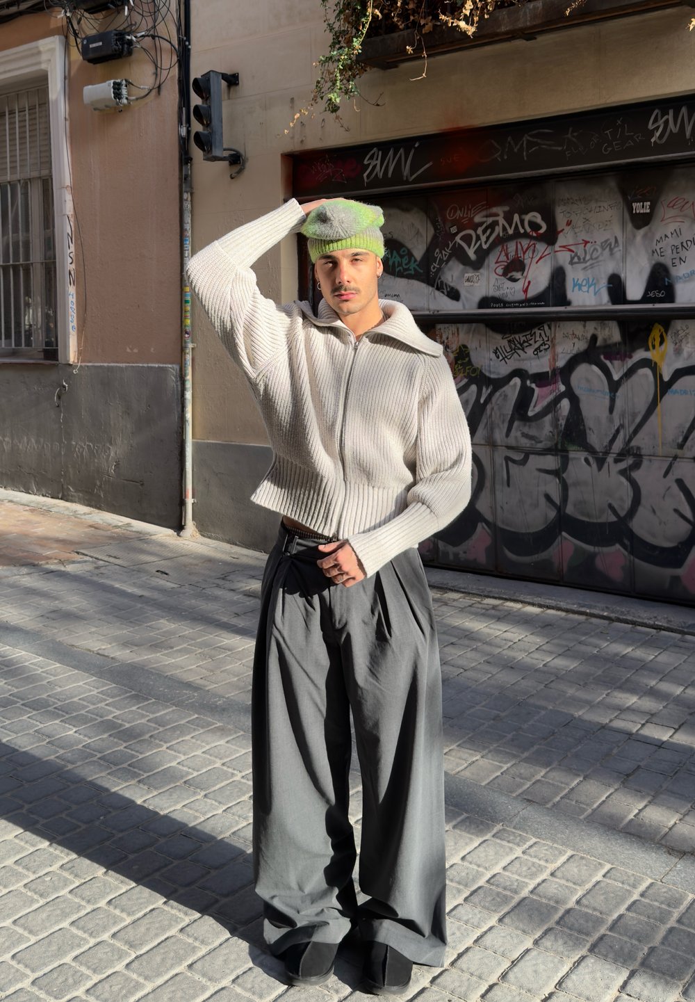 Jeune personne portant un pull en tricot blanc, un pantalon gris à jambes larges et un bonnet vert, debout dans une rue urbaine ensoleillée avec des graffitis en arrière-plan.