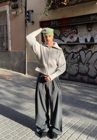 Jeune personne portant un pull en tricot blanc, un pantalon gris à jambes larges et un bonnet vert, debout dans une rue urbaine ensoleillée avec des graffitis en arrière-plan.