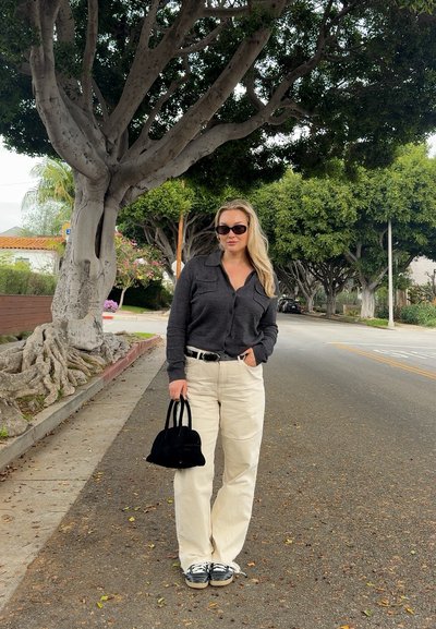 Mujer con gafas de sol, camisa oscura de manga larga y pantalones claros, de pie en una calle arbolada sosteniendo un bolso negro.