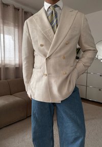 Άνδρας που φοράει μπεζ διπλόστενη σακάκι, λευκό πουκάμισο, γραβάτα με ρίγες και μπλε τζιν, στέκεται μέσα σε εσωτερικό χώρο με το ένα χέρι στην τσέπη.